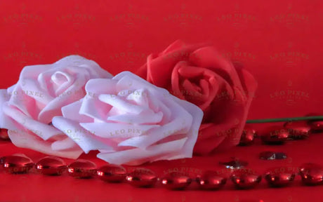 Three roses, two in soft pink and one in deep red, rest against a red background. Small red heart-shaped gems are scattered around, reflecting light. The petals have a velvety texture, and the green stem of the red rose adds contrast. The smooth backdrop enhances the warm tones, while the gems create a delicate sparkle in the composition. Photography.