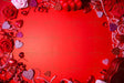 A red background is decorated with roses in pink and red, sparkling glitter hearts, and glossy heart-shaped ornaments. The frame is filled with shredded paper, small heart chocolates, and a red heart-shaped box. The center is left empty, creating a balanced composition. The bright lighting highlights the rich textures and colors, enhancing the festive and romantic theme. Photography.