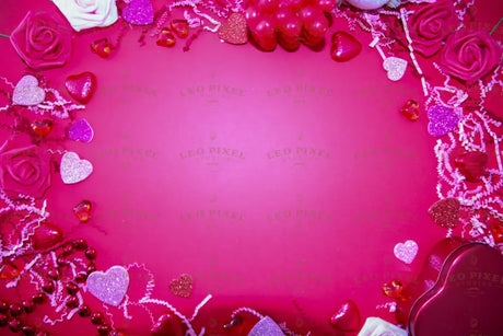 A pink background is decorated with roses in pink and red, sparkling glitter hearts, and glossy heart-shaped ornaments. The frame is filled with shredded paper, small heart chocolates, and a red heart-shaped box. The center is left empty, creating a balanced composition. The bright lighting highlights the rich textures and colors, enhancing the festive and romantic theme. Photography