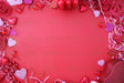 A red background is decorated with roses in pink and red, sparkling glitter hearts, and glossy heart-shaped ornaments. The frame is filled with shredded paper, small heart chocolates, and a red heart-shaped box. The center is left empty, creating a balanced composition. The bright lighting highlights the rich textures and colors, enhancing the festive and romantic theme. Photography.