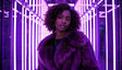 A stylish woman stands confidently in a hallway illuminated by vivid purple neon lights. She wears a luxurious purple fur coat, her curly hair framing her face beautifully. Her expression is poised and serene, accented by delicate hoop earrings and a pendant necklace. The glowing vertical neon lights create a futuristic and dramatic ambiance, enhancing the overall elegance of the image. Ai generated. Photography style.