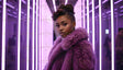 A poised woman in a deep violet fur coat stands in a futuristic setting with glowing vertical neon lights in soft purple hues. Her styled hair adds sophistication, complementing her elegant expression. The vivid lighting creates striking reflections on the glass panels around her, adding depth and modernity to the composition, while subtle background figures enhance the dynamic ambiance. Ai generated. Photography style.