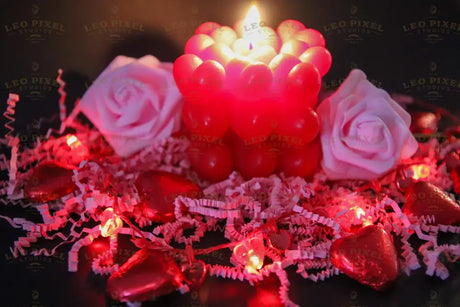 A red candle made of small heart shapes burns softly, casting a warm glow. Two pink roses rest on crinkled pink paper shreds. Red heart-shaped chocolates and heart-shaped fairy lights decorate the scene. The dark background creates contrast, making the reds and pinks stand out beautifully. The soft lighting and shallow depth of field add warmth and depth to the image. Photography.