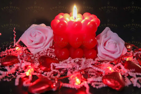 A red candle made of small heart shapes burns softly, casting a warm glow. Two pink roses rest on crinkled pink paper shreds. Red heart-shaped chocolates and heart-shaped fairy lights decorate the scene. The dark background creates contrast, making the reds and pinks stand out beautifully. The soft lighting and shallow depth of field add warmth and depth to the image. Photography.