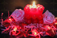 A red candle made of small heart shapes burns softly, casting a warm glow. Two pink roses rest on crinkled pink paper shreds. Red heart-shaped chocolates and heart-shaped fairy lights decorate the scene. The dark background creates contrast, making the reds and pinks stand out beautifully. The soft lighting and shallow depth of field add warmth and depth to the image. Photography.