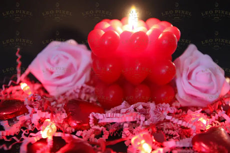 A red candle made of small heart shapes burns softly, casting a warm glow. Two pink roses rest on crinkled pink paper shreds. Red heart-shaped chocolates and heart-shaped fairy lights decorate the scene. The dark background creates contrast, making the reds and pinks stand out beautifully. The soft lighting and shallow depth of field add warmth and depth to the image. Photography. 