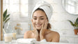 A smiling woman with a towel wrapped around her head gently touches her face in a bright, modern bathroom. The space features a white countertop with neatly placed items, including a pump bottle, a cup, and a folded towel. A round mirror, leafy plant, and soft natural lighting add to the clean and refreshing ambiance of the setting. The gold faucet adds a subtle touch of elegance. Ai generated. Photography style.